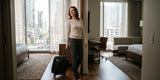 Woman wearing dark wide-leg pants and a beige long-sleeve top, pulling a black carry-on suitcase through a hotel room with city views.