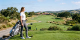 Woman wearing dark gray golf pants, a white polo shirt, and white shoes, pulling a golf trolley across a tee box overlooking a sunny golf course.
