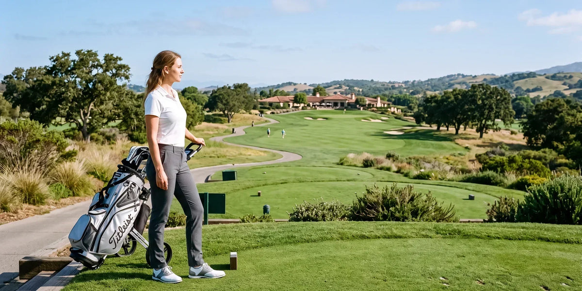 Woman wearing dark gray golf pants, a white polo shirt, and white shoes, pulling a golf trolley across a tee box overlooking a sunny golf course.