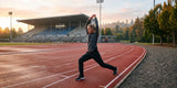 Woman wearing black track pants and a dark gray long-sleeve top, stretching in a lunge with arms overhead on an outdoor running track at sunset.