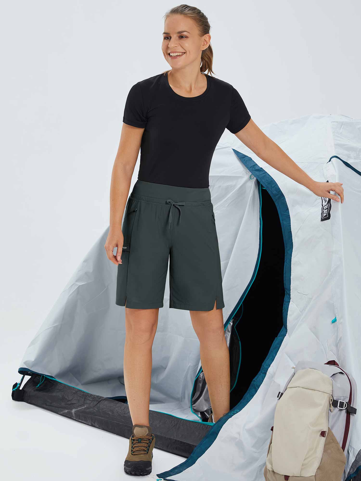 Woman in dark gray quick-dry hiking Bermuda shorts standing by a tent. #color_Dark Gray